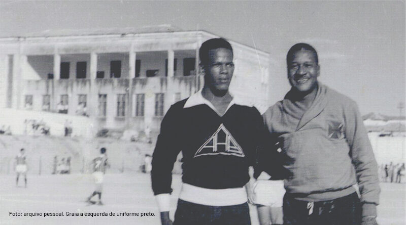 Na imagem: dois homens, em frente a um campo de futebol, o homem do lado esquerdo veste uniforme preto e o do lado direito uniforme branco. A foto está em preto e branco.